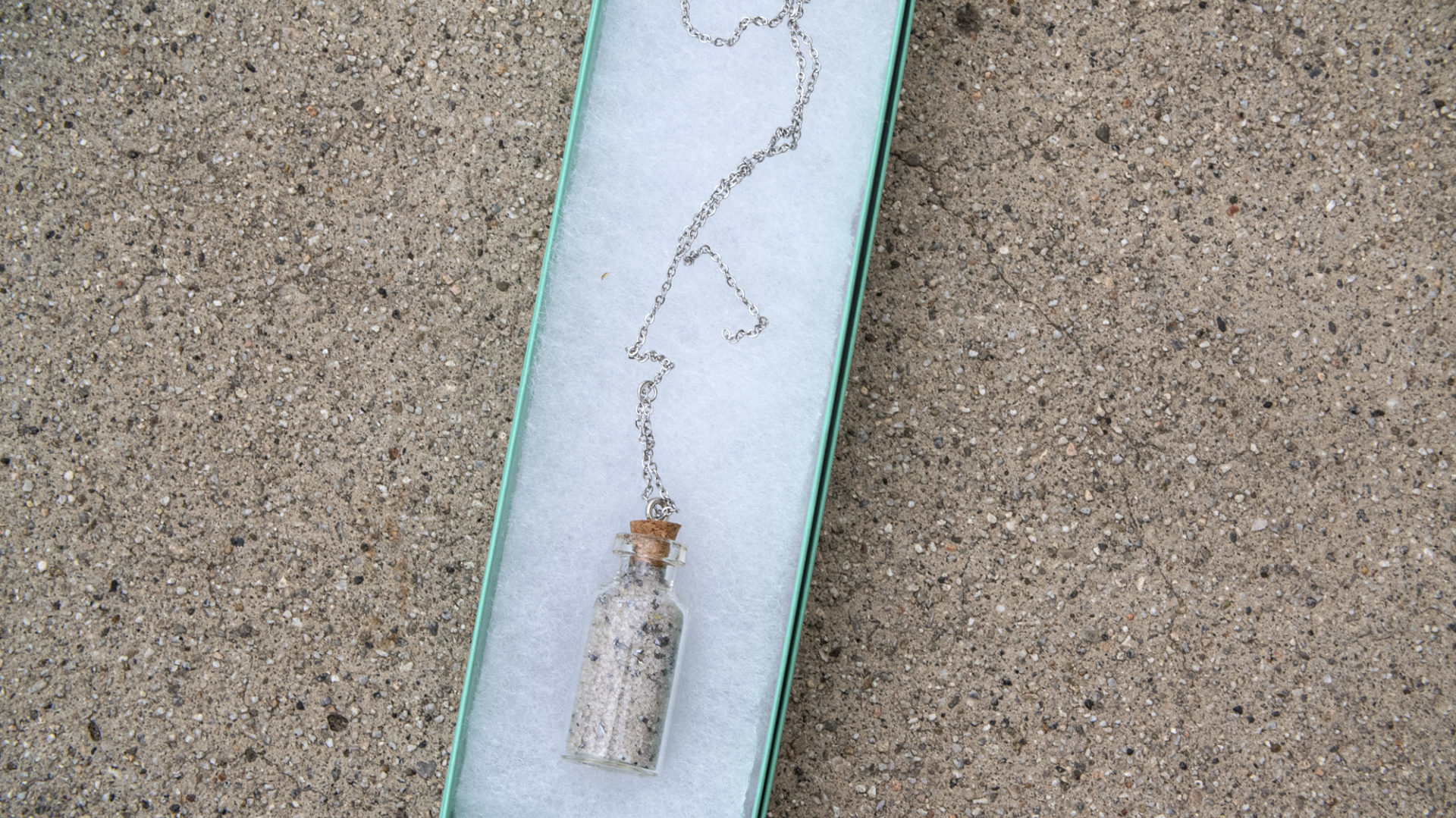 A small glass souvenir bottle filled with sand and bits of metal, on a silver chain, in a sea-foam green box, over concrete.